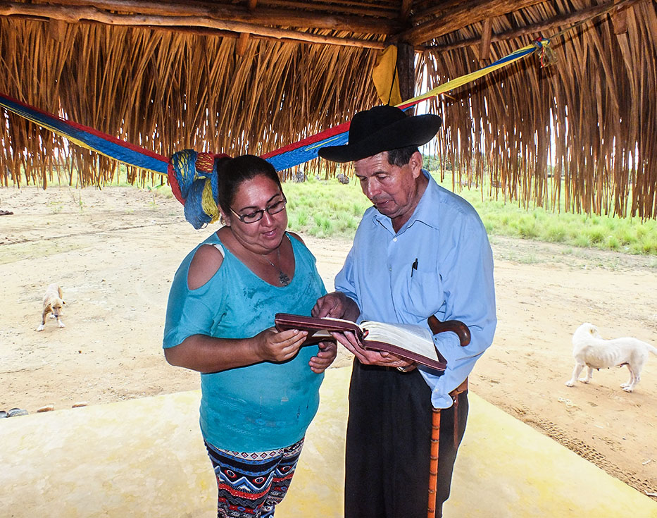 Colombian Christian missionary reads the Bible to a woman under a grass-roofed pavilion