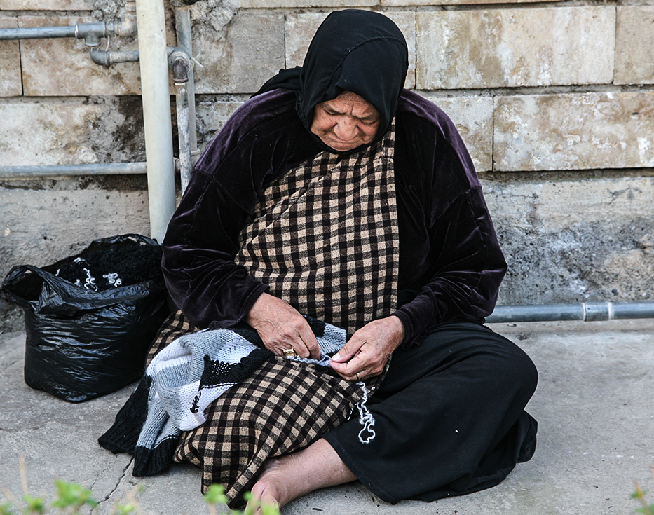 Iraqi woman sits on a concrete sidewalk pulling apart knitted scarf
