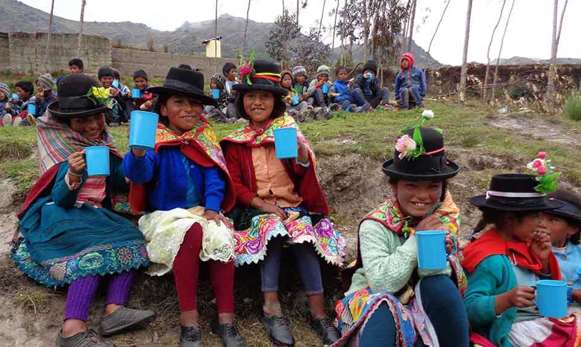 Peruvian girls wearing decorative dresses sit on the ground drinking water from blue mugs