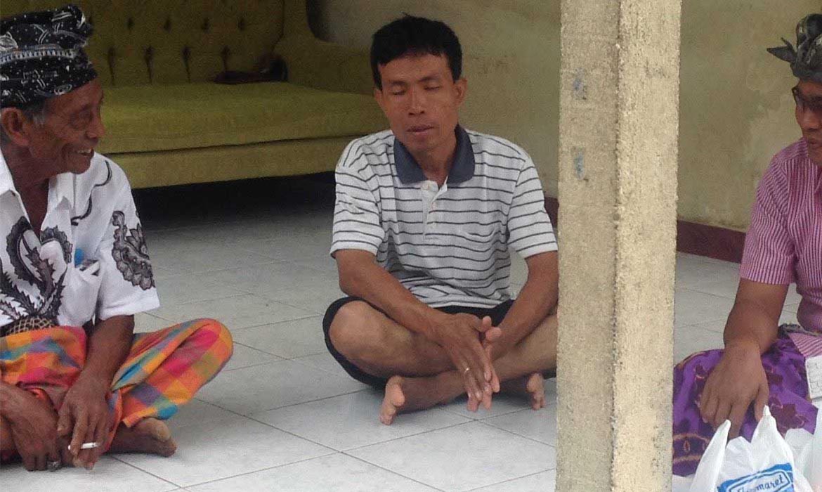 Southeast Asian man sits on tile floor with legs crossed and hands together while others sit in a circle with him