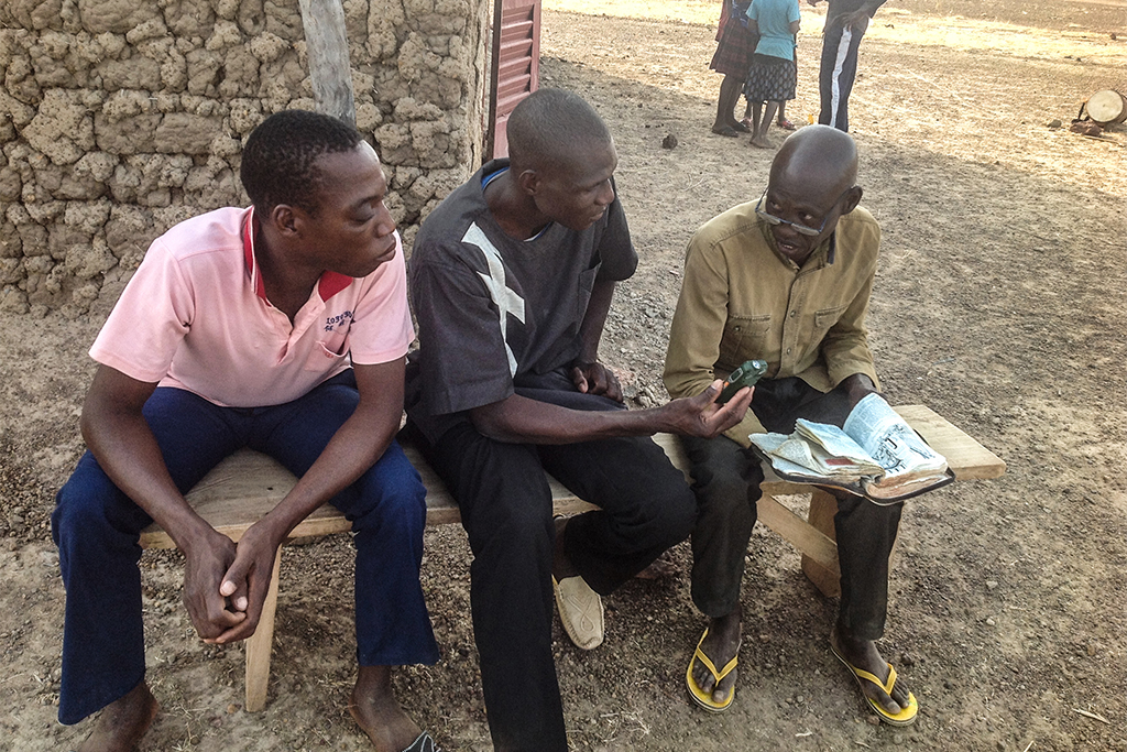 Two men one holding a phone sit on a bench listening to a Burkinabé Christian missionary teaching from his Bible