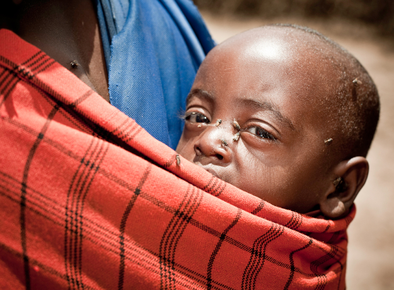 African boy resting in a sling around his mother's shoulder with flies flying around his face