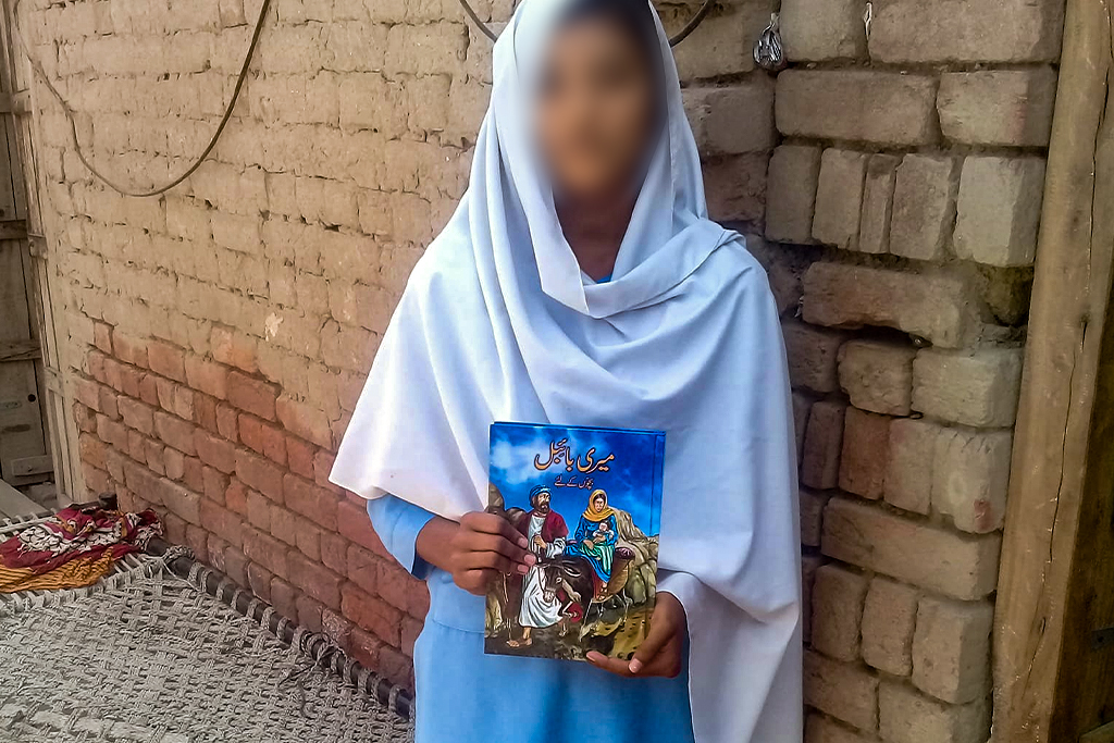 Pakistani Christian girl standing in front of a brick wall holding illustrated Bible