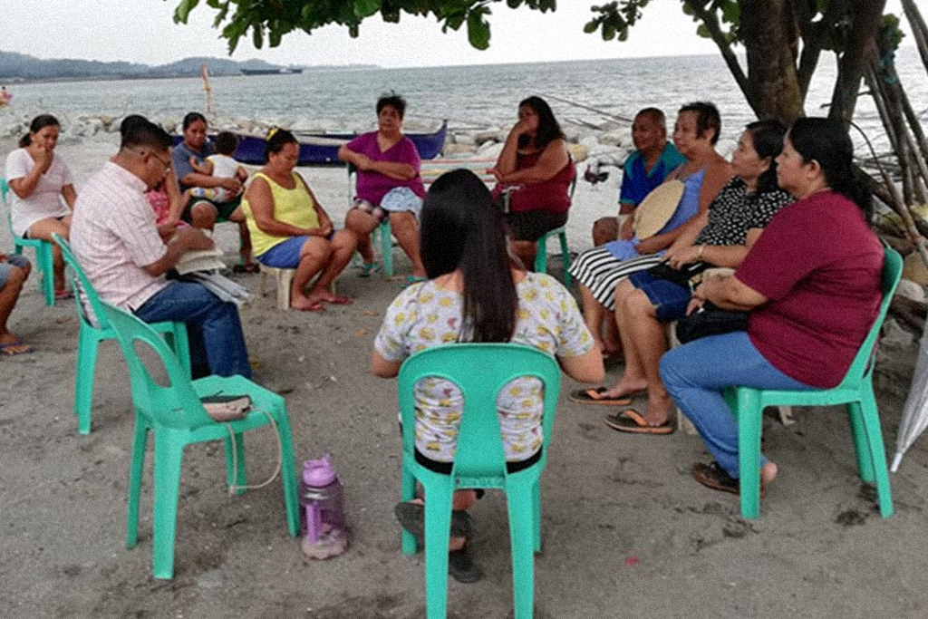 Filipino Christians sitting in plastic teal lawn chairs under a tree on a beach
