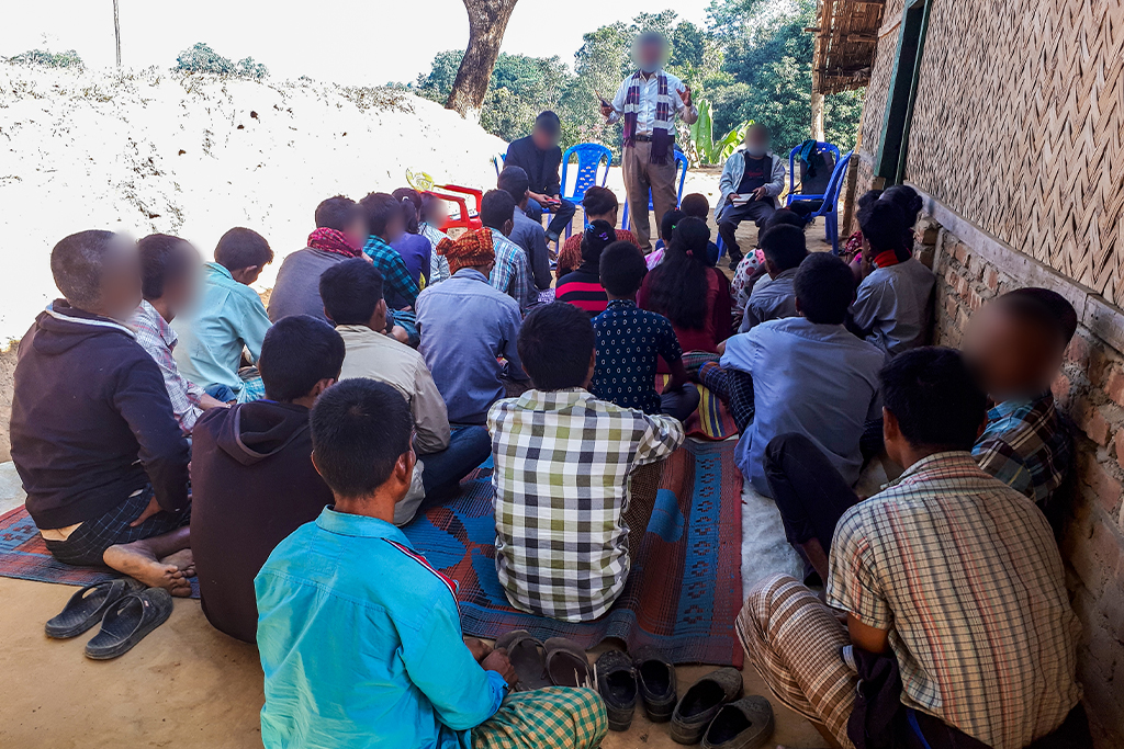 South Asian Christian missionary preaches the gospel to young men and women sitting outside on dry ground next to a house