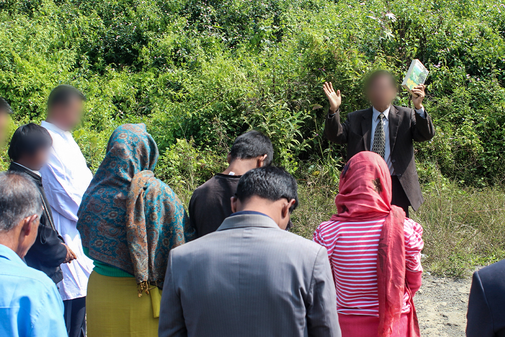 South Asian Christian missionary wearing a brown suit and holding a Bible proclaims the gospel to a group of men and women