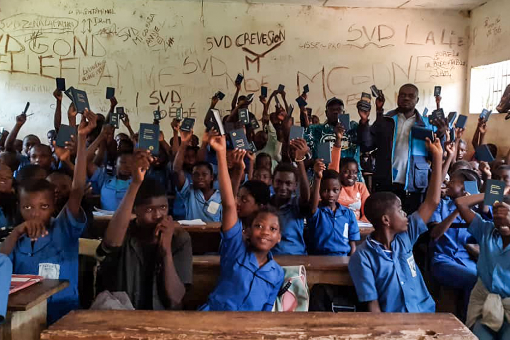 School children in Cameroon wearing blue uniforms sitting at wooden desks holding up small books