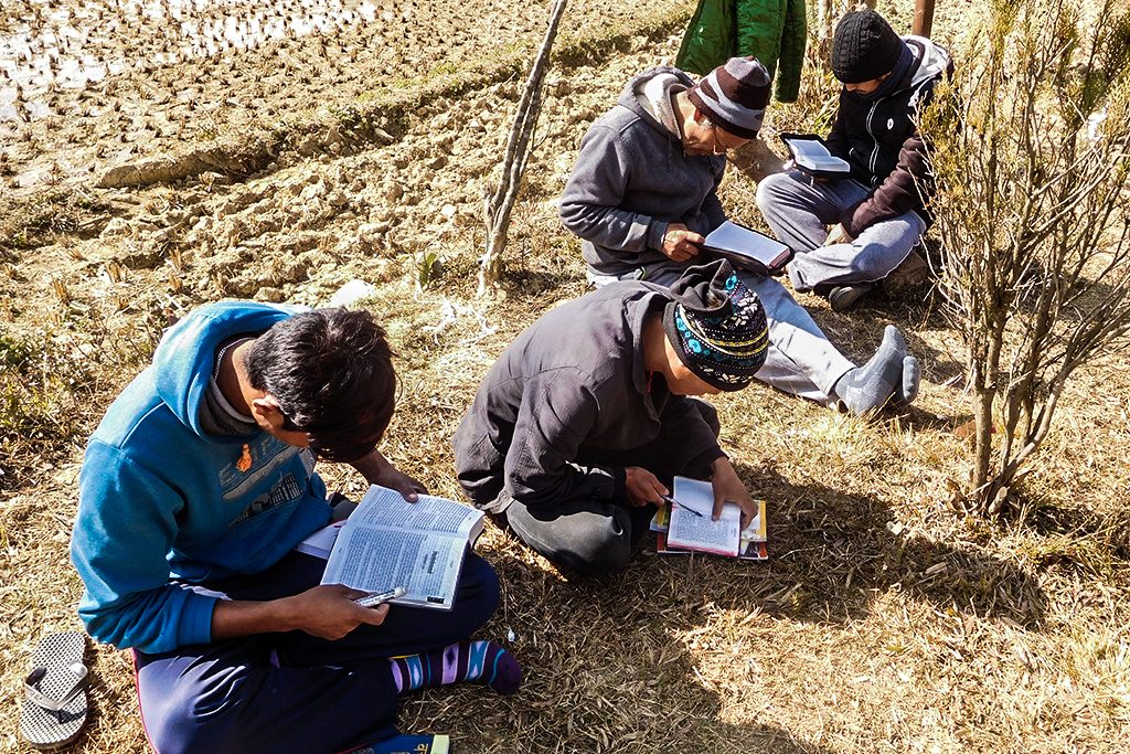 Nepali Christians wearing sweatshirts and sweatpants sitting in a barren field reading their Bibles