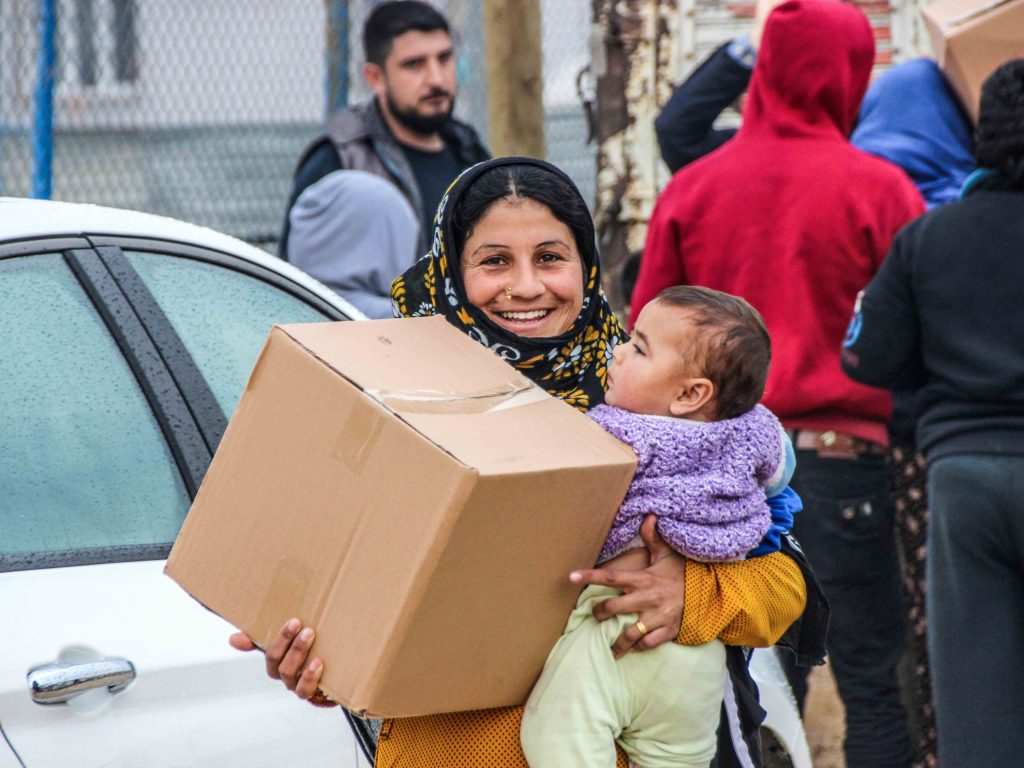 A refugee woman holding her baby in one arm and a box of goods in the other smiling