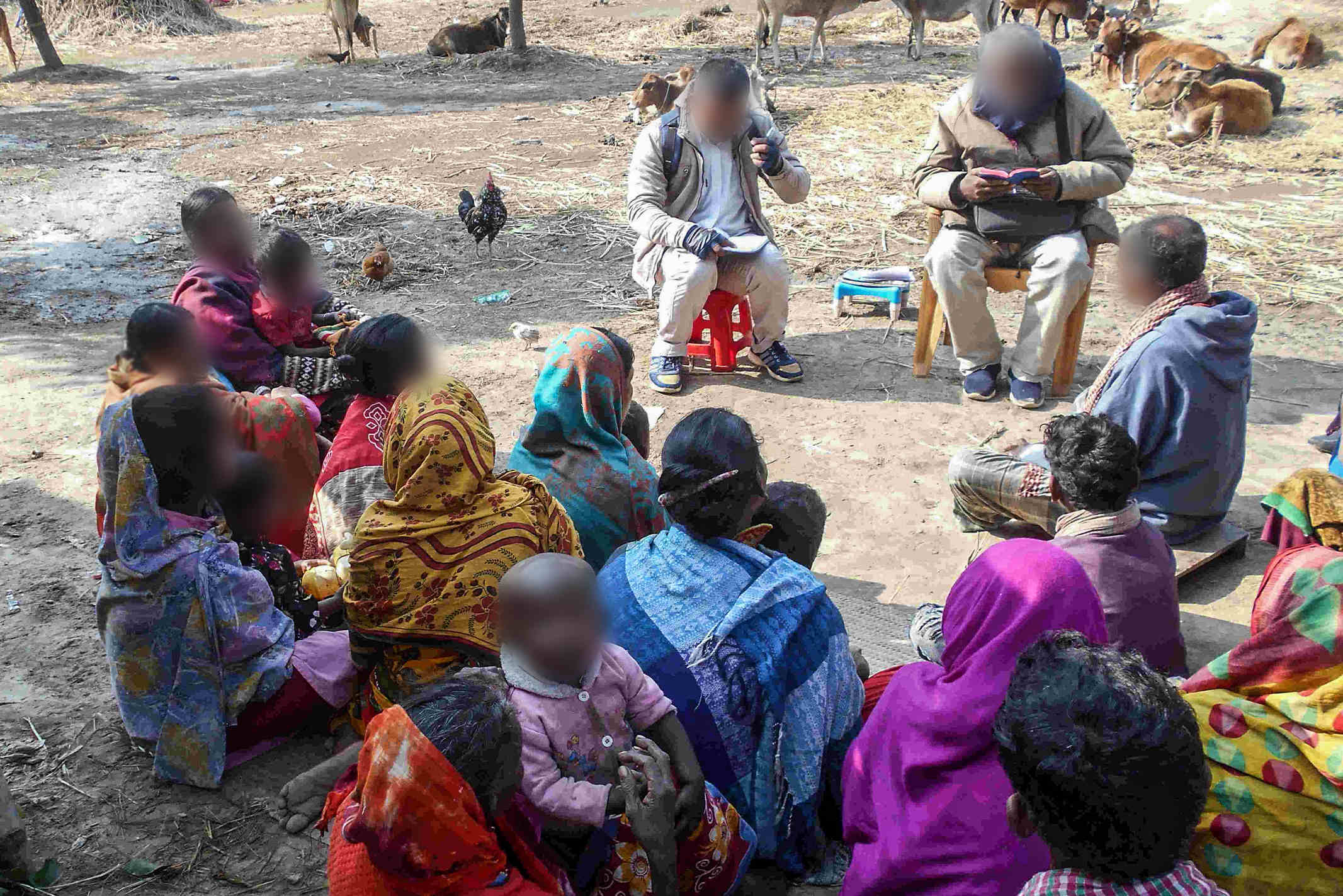 Christian missionaries sitting outside on plastic stools and chairs read the Bible to a group of Bangledeshi woman and children