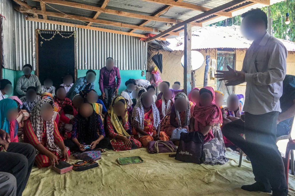 A missionary in South Asia shares the gospel to a group of men and women wearing formal clothing on a porch covered by a tin roof