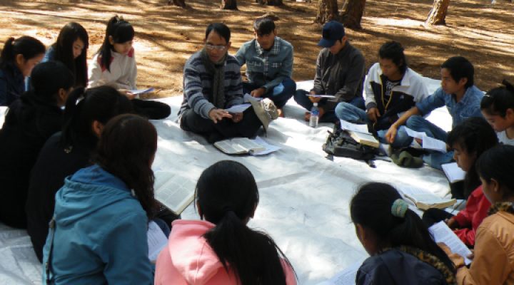 Christian missionary leads Bible study of young adult Christians who sit in a circle on the ground